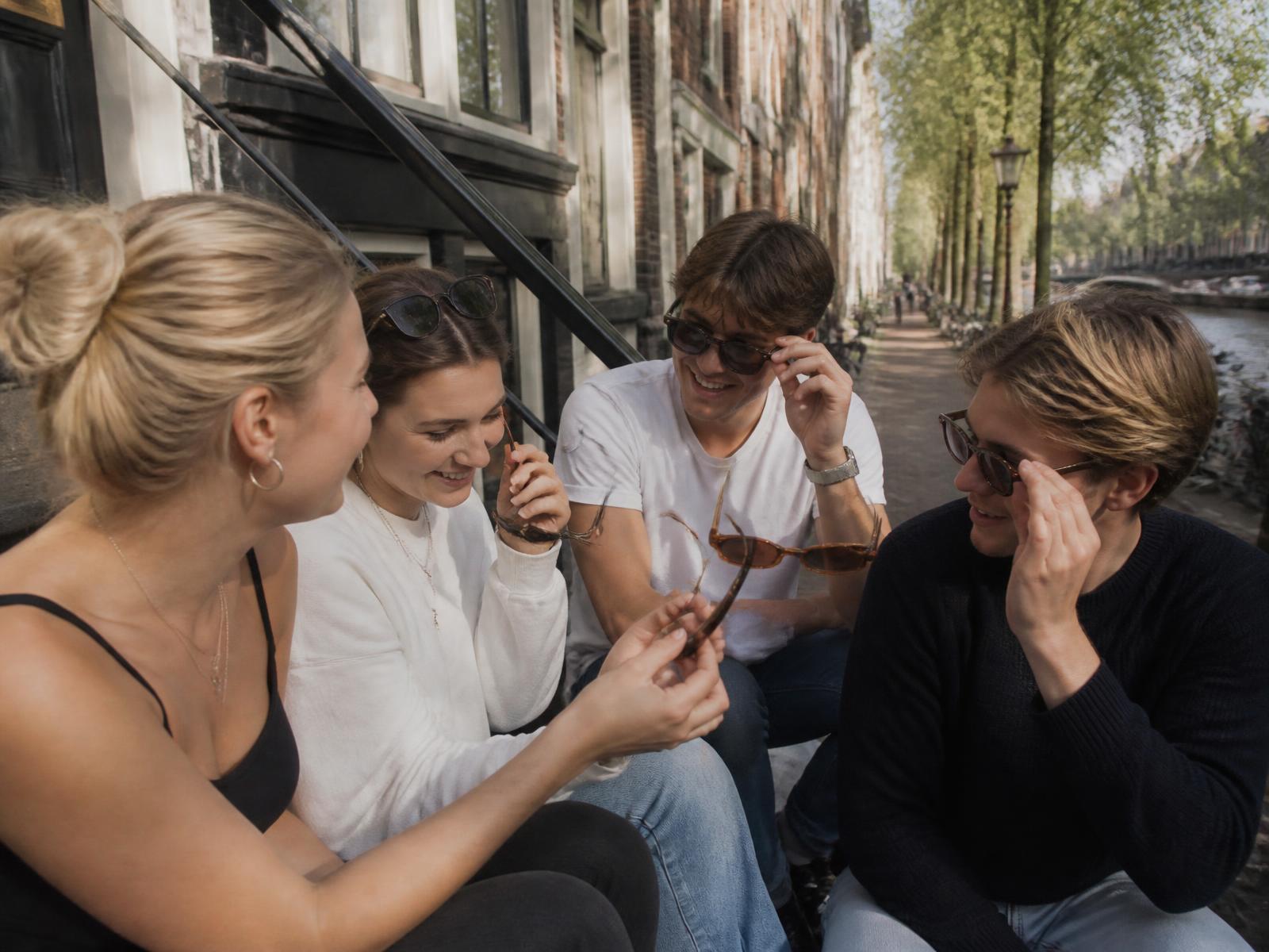 Vrienden passen samen zonnebrillen op een stoep langs een Amsterdamse gracht